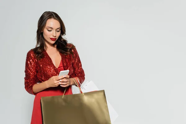 Elegante donna bruna in abito rosso utilizzando smartphone e tenendo le borse della spesa isolate sul grigio — Foto stock