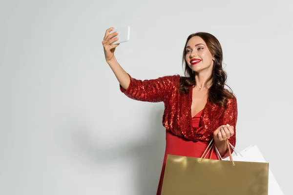 Mujer morena sonriente en vestido rojo tomando selfie en el teléfono inteligente y sosteniendo bolsas de compras sobre fondo gris - foto de stock