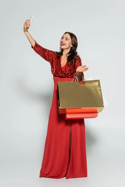 Pleine longueur de femme gaie en robe élégante prenant selfie et tenant des sacs à provisions sur fond gris — Photo de stock
