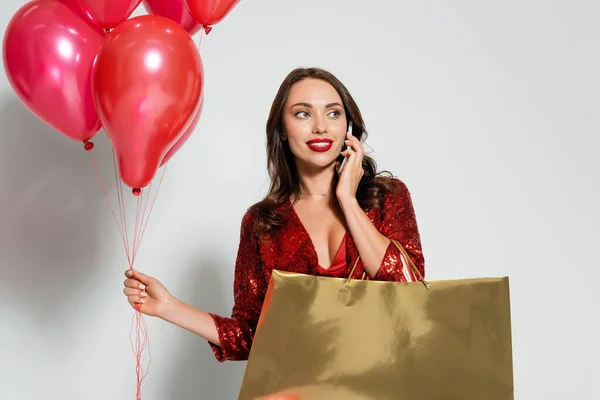 Smiling woman with red lips talking on smartphone and holding shopping bags and balloons on grey background — Stock Photo