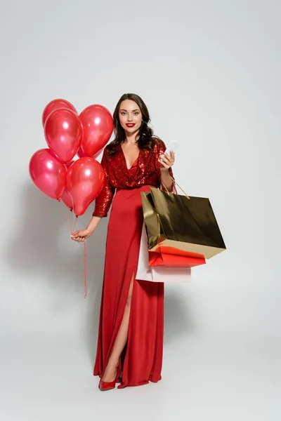 Full length of woman in red dress holding smartphone and balloons on grey background — Stock Photo