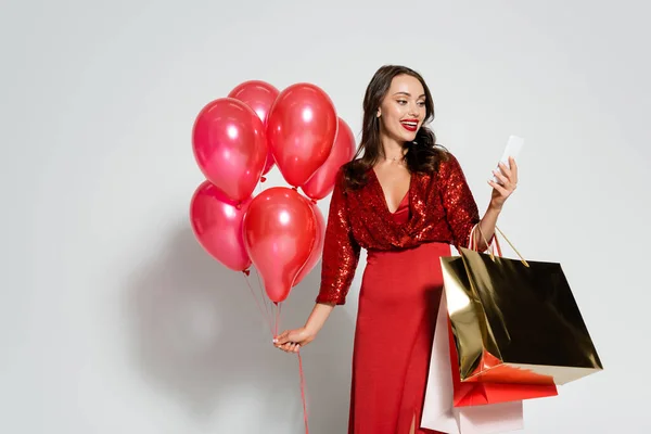 Веселая брюнетка в красном платье с помощью смартфона и держа сумки с шариками на сером фоне — стоковое фото