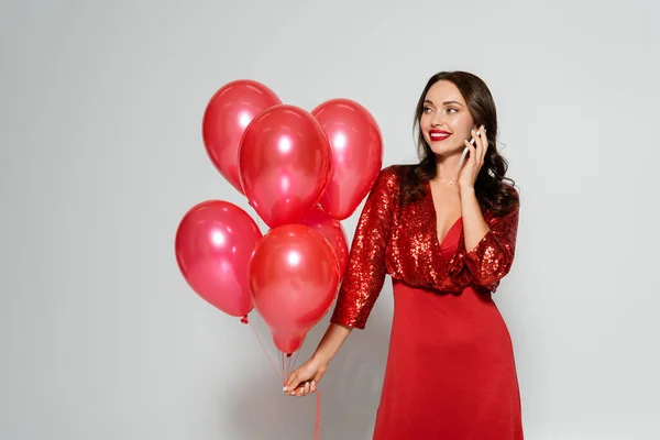 Mujer alegre en vestido rojo elegante hablando en el teléfono celular y sosteniendo globos sobre fondo gris - foto de stock