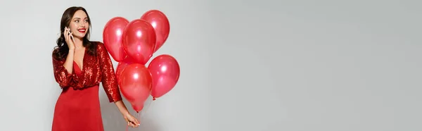 Hübsche Frau in rotem Kleid hält Luftballons in der Hand und spricht auf Smartphone auf grauem Hintergrund, Banner — Stockfoto