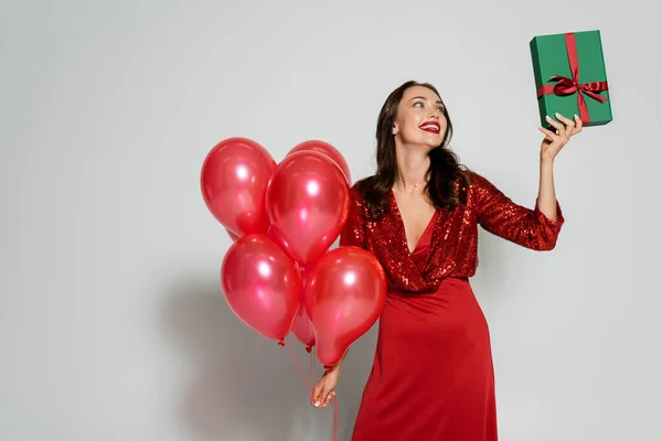 Весела жінка в червоній сукні тримає подарунок і повітряні кулі на сірому фоні — стокове фото