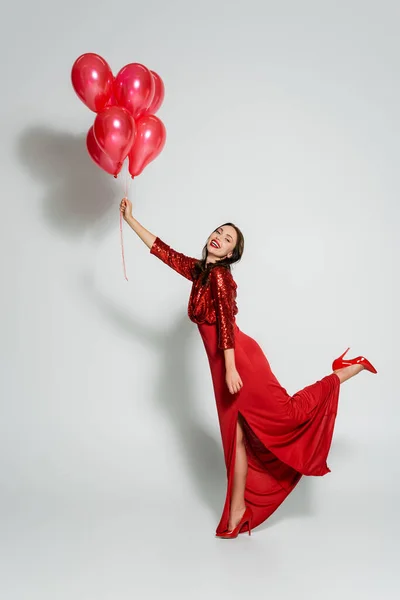 Allegro giovane donna in abito rosso che tiene palloncini e guardando la fotocamera su sfondo grigio — Foto stock