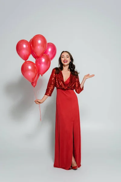 Full length of pretty woman in red dress holding balloons and pointing with hand on grey background — Stock Photo