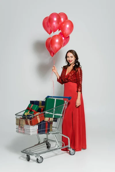 Mujer bastante joven en vestido elegante sosteniendo globos cerca del carrito de compras con regalos sobre fondo gris - foto de stock