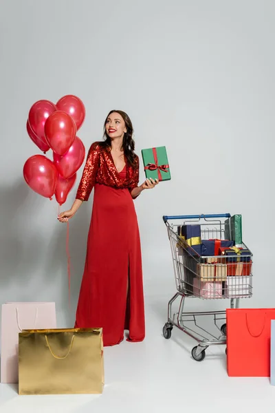 Mujer alegre elegante sosteniendo globos y presente cerca del carrito de la compra y bolsas sobre fondo gris - foto de stock