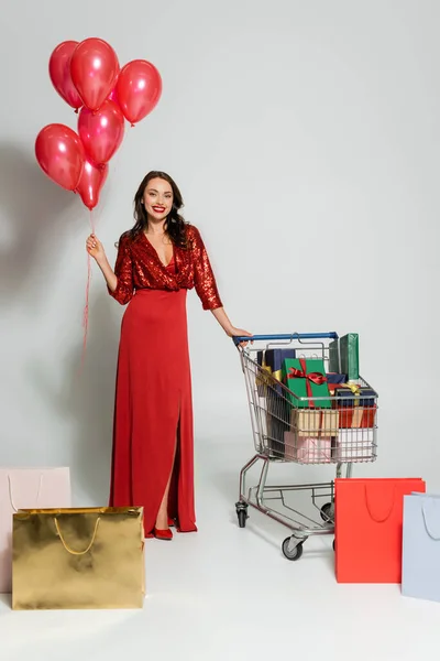 Mujer alegre en vestido sosteniendo globos cerca del carrito de la compra con regalos y compras sobre fondo gris - foto de stock