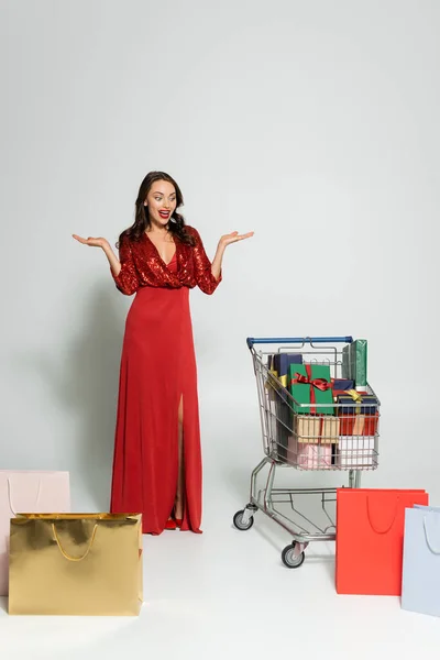 Mulher excitada em vestido vermelho olhando para carrinho com presentes e sacos de compras em fundo cinza — Fotografia de Stock