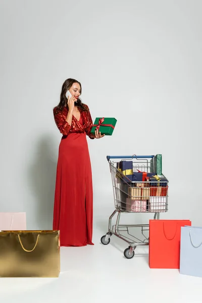 Mulher elegante falando no smartphone e segurando presente perto de sacos de compras no fundo cinza — Fotografia de Stock