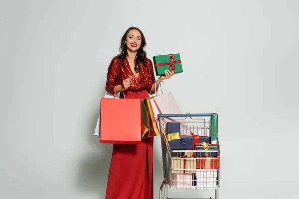 Mujer alegre en vestido elegante sosteniendo bolsas de compras y regalo sobre fondo gris - foto de stock