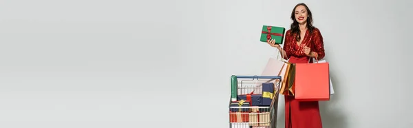 Cheerful young woman in dress holding present and shopping bags near cart on grey background, banner — Stock Photo