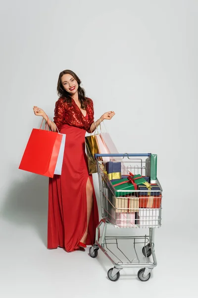 Donna positiva in abito rosso con borse della spesa vicino al carrello con regali su sfondo grigio — Foto stock