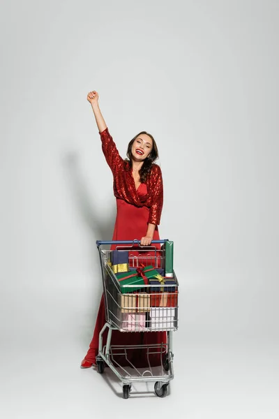 Mujer excitada en vestido rojo de pie cerca del carrito de la compra con regalos sobre fondo gris - foto de stock