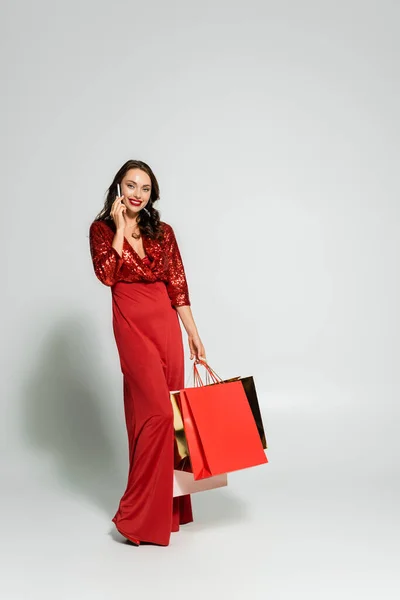 Stylish woman in dress talking on smartphone and holding shopping bags on grey background — Stock Photo