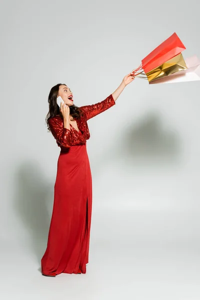 Mujer excitada en vestido rojo hablando en el teléfono inteligente y sosteniendo bolsas de compras sobre fondo gris - foto de stock