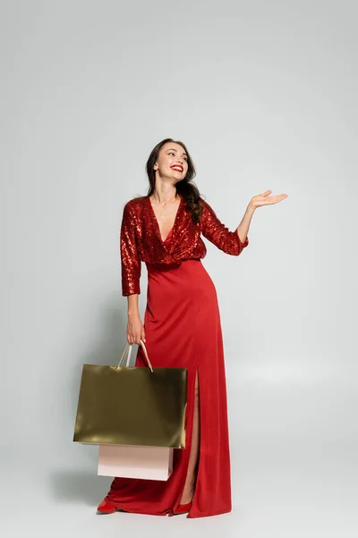 Happy woman in red dress pointing with hand and holding shopping bags on grey background — Stock Photo