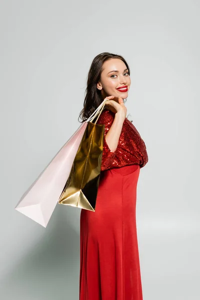 Mujer alegre en vestido rojo festivo sosteniendo bolsas de compras sobre fondo gris - foto de stock