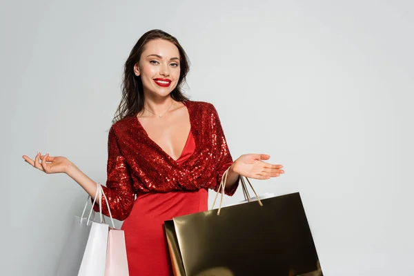 Mulher morena feliz em vestido vermelho olhando para a câmera enquanto segurando sacos de compras isolados em cinza — Fotografia de Stock