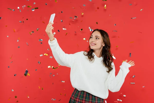 Sonriente morena tomando selfie en smartphone bajo confeti sobre fondo rojo - foto de stock