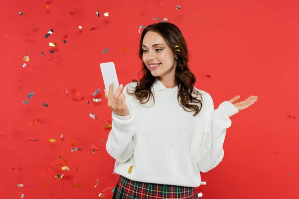 Fröhliche Frau im kuscheligen Pullover mit Smartphone unter festlichem Konfetti auf rotem Hintergrund — Stockfoto