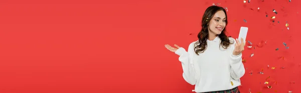 Положительная брюнетка женщина указывая рукой и используя смартфон под конфетти на красном фоне, баннер — стоковое фото