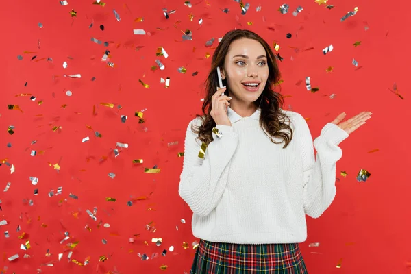 Aufgeregte Frau im Pullover unterhält sich auf Smartphone unter Konfetti auf rotem Hintergrund — Stockfoto