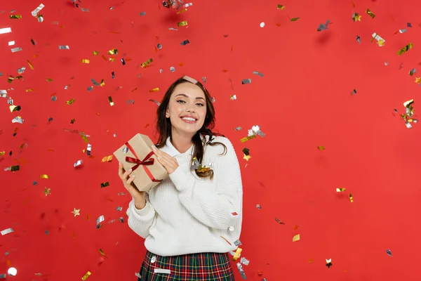 Весела брюнетка в светрі тримає подарунок під конфетті на червоному тлі — стокове фото
