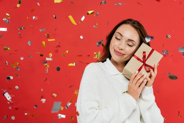Улыбающаяся женщина в тёплом свитере держит подарок с бантиком под конфетти на красном фоне — стоковое фото