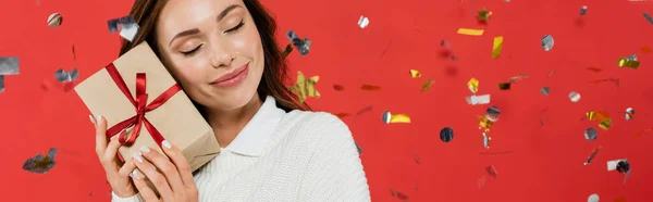 Mujer morena sonriente en suéter sosteniendo presente bajo confeti sobre fondo rojo, pancarta - foto de stock