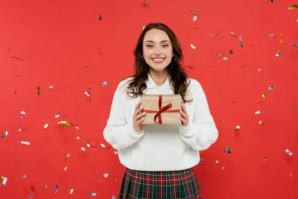 Fröhliche brünette Frau im warmen Pullover hält Geschenk mit Schleife unter Konfetti auf rotem Hintergrund — Stockfoto