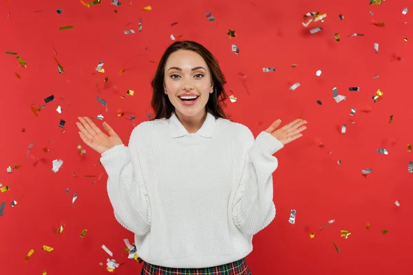 Excited brunette woman in warm sweater looking at camera under confetti on red background — Stock Photo