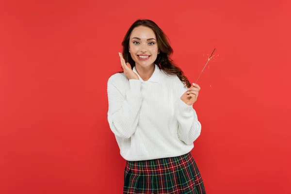 Positive brunette woman holding sparkler and looking at camera isolated on red — Stock Photo
