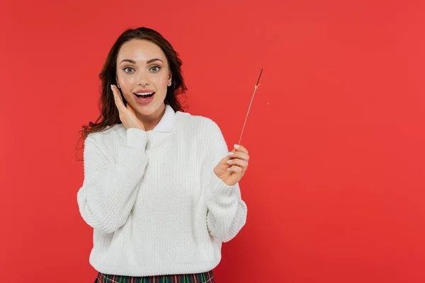 Excited woman in warm sweater holding sparkler and looking at camera isolated on red — Stock Photo