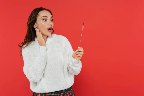 Shocked woman in sweater looking at sparkler isolated on red — Stock Photo