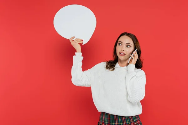 Brunette woman in sweater holding speech bubble and talking on smartphone on red background — Stock Photo