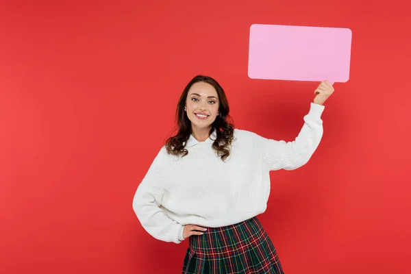 Positive brunette woman in sweater holding empty speech bubble on red background — Stock Photo