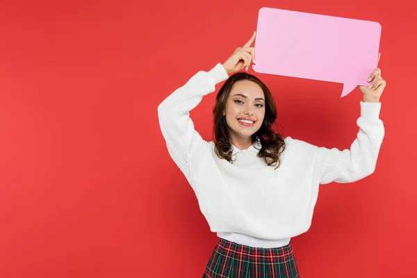 Positive brunette woman holding speech bubble and looking at camera on red background — Stock Photo
