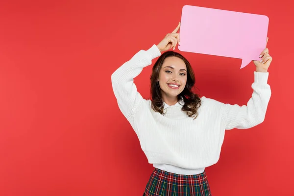 Mulher morena positiva segurando bolha de fala e olhando para a câmera no fundo vermelho — Fotografia de Stock