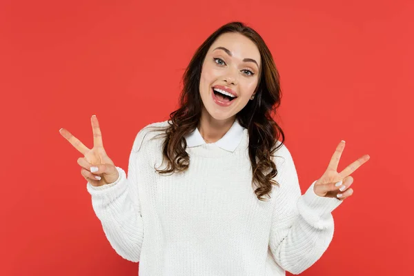 Positive brunette woman in white jumper showing peace sign isolated on red — Stock Photo