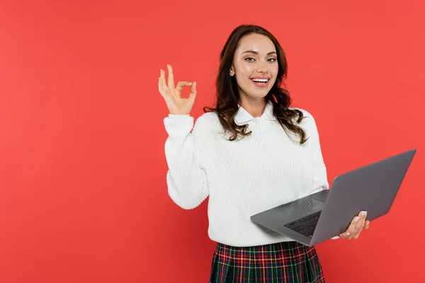 Femme souriante en pull confortable montrant un geste correct et tenant l'ordinateur portable isolé sur rouge — Photo de stock