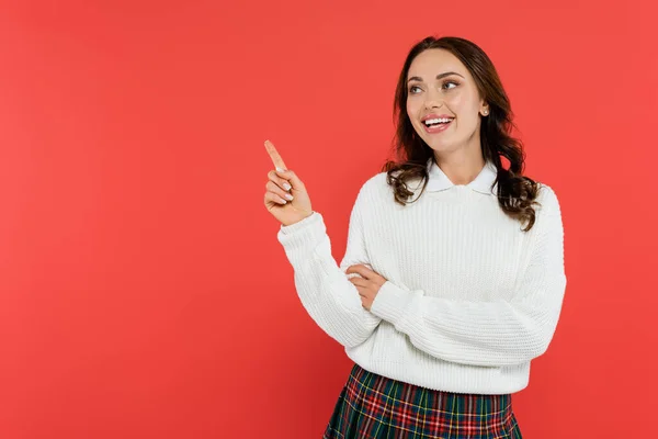 Cheerful brunette woman in jumper pointing with finger isolated on red — Stock Photo