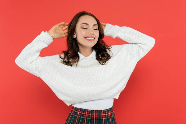 Cheerful woman in warm jumper touching hair isolated on red — Stock Photo