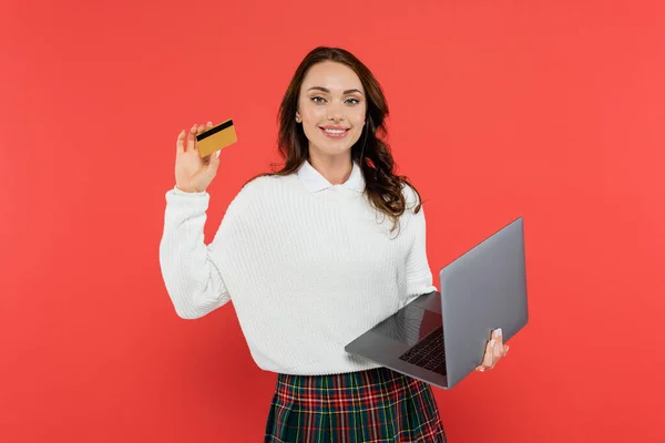 Mujer sonriente en jersey con tarjeta de crédito y portátil aislado en rojo - foto de stock