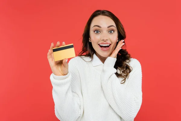 Aufgeregt brünette Frau im weißen Pullover hält Kreditkarte isoliert auf rot — Stockfoto