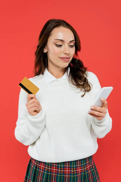 Mujer sonriente en jersey con tarjeta de crédito y el uso de teléfono inteligente aislado en rojo - foto de stock