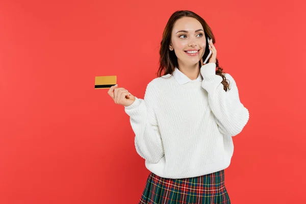 Cheerful young woman in jumper talking on smartphone and holding credit card isolated on red — Stock Photo