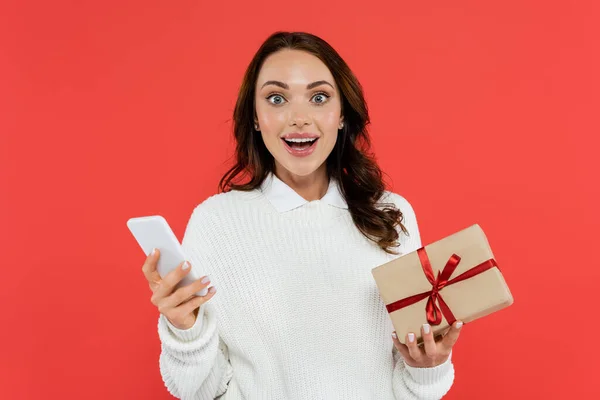 Aufgeregte Frau im Pullover hält Smartphone und Geschenk auf Rot — Stockfoto
